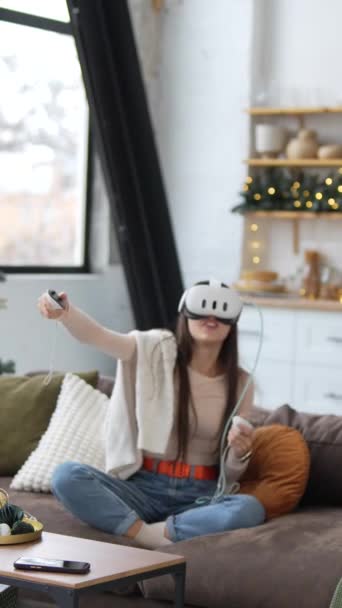 Temidden van de kerstboom achtergrond, een jonge en mooie dame geniet van gaming via een virtual reality headset terwijl u op de bank zit. Hoge kwaliteit 4k beeldmateriaal - Video
