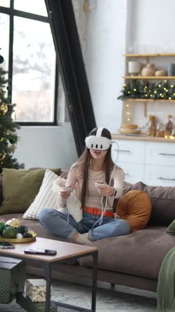 Mladá a krásná dáma sedí na pohovce s vánoční stromeček v pozadí, hraní přes virtuální realitu sluchátka. Vysoce kvalitní 4K záběry - Záběry, video