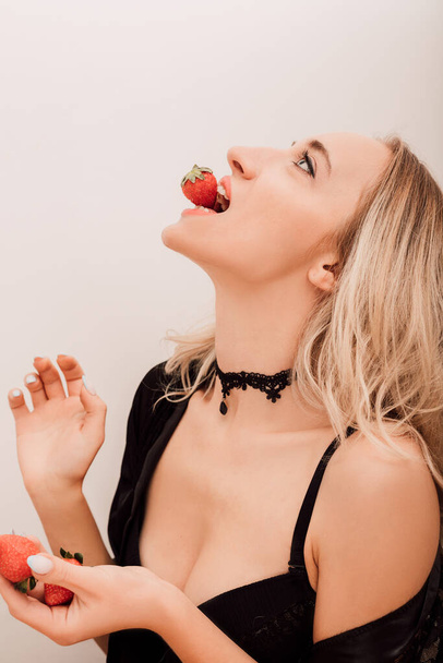 A nő epret eszik. Szexi szőke nő fekete fürdőköpenyben a konyhában bogyókat eszik. Kiváló minőségű fénykép - Fotó, kép