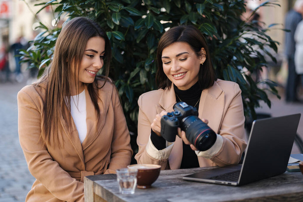 Female photographer checking pictures in camera with her client. Women sitting in cafe and drinking coffee. Work space outdoors. High quality photo - Photo, Image