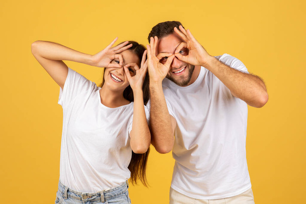 Alegre joven europeo hombre y mujer en camisetas blancas haciendo gestos de gafas divertidas con las manos, disfrutar del tiempo libre, aislado en el fondo del estudio amarillo. Emociones y estilo de vida - Foto, Imagen