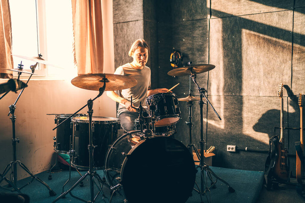 Ο ντράμερ παίζει ντραμς στο στούντιο μουσικής στην πρόβα της μπάντας. Μουσικός με όργανο στο όμορφο φως ηλιοβασίλεμα από το παράθυρο. - Φωτογραφία, εικόνα