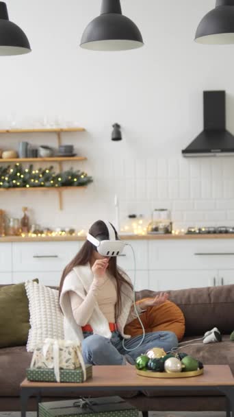 Una joven y atractiva dama se sienta junto al árbol de Navidad, jugando videojuegos con la ayuda de un auricular de realidad virtual. Imágenes de alta calidad 4k - Metraje, vídeo