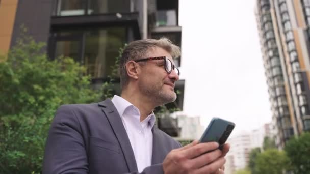 Onnellinen liikemies laseissa käyttäen älypuhelinta ulkona kaupungissa. Liiketoiminta, viestintä, teknologia. Reaaliaikainen käsite - Materiaali, video