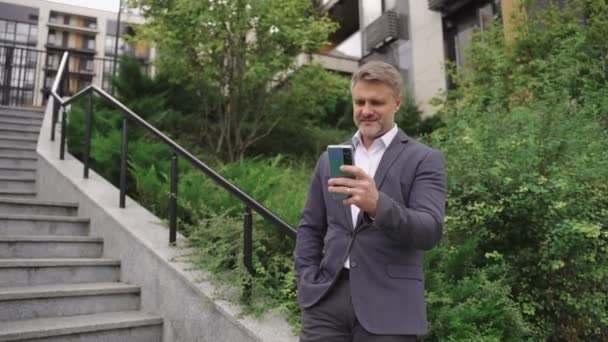 Усміхнений бізнесмен, використовуючи смартфон, стоїть біля сходів у місті. Бізнес, комунікація, концепція технології. Концепція реального часу - Кадри, відео