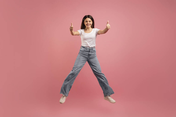 Радостная девочка-подросток прыгает в воздухе и снимает пальцы на камеру, а также веселится на розовом студийном фоне, в полный рост, в пространстве для копирования. - Фото, изображение