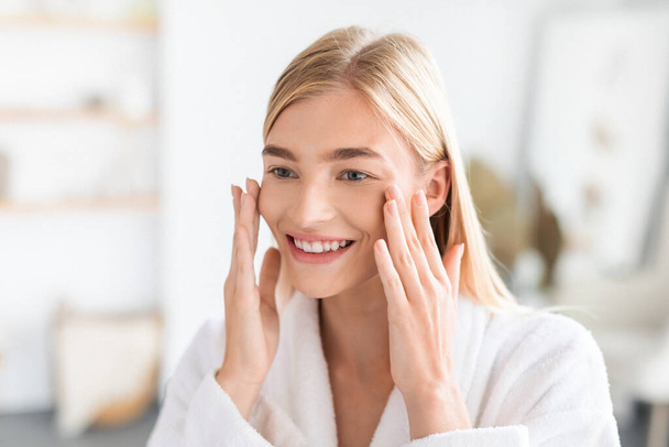 Blonde lady in white robe enjoys freshness of her skin touching face with smile, enjoying result of new skincare routine, standing in bathroom at home. Beauty and pampering, cosmetics - Foto, afbeelding