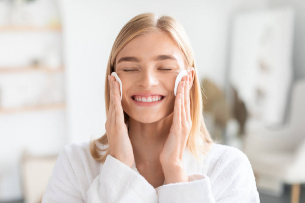 Young blonde lady smiles applying tonic with cotton pads, pressing them to cheeks, standing with closed eyes in modern bathroom in the morning, embodying fresh facial skincare routine - Fotoğraf, Görsel