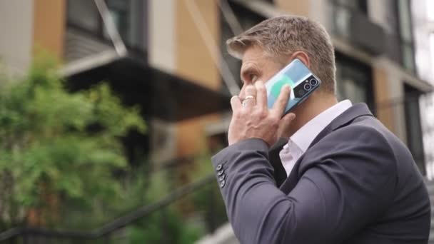 Усміхнений стильний дорослий чоловік розмовляє по смартфону, стоїть надворі. Бізнес, комунікація, концепція технології. Концепція реального часу - Кадри, відео