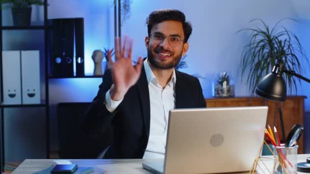Hello. Indian business man working on laptop computer smiling friendly at camera and waving hands gesturing hi, greeting or goodbye, welcoming with hospitable expression at home office workplace desk - Footage, Video