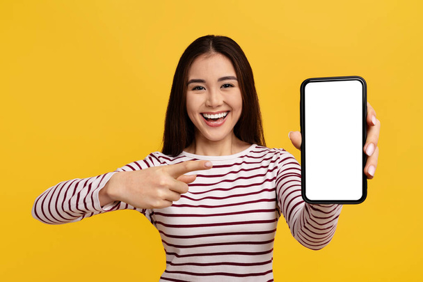 Pozitif Çinli 20 'li yaşlarda genç bir bayan beyaz ekranlı ve gülümseyen büyük bir telefon gösteriyor, güzel mobil uygulamalar öneriyor, harika bir online teklif, sarı arka planda izole edilmiş. - Fotoğraf, Görsel