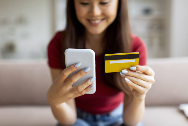 Zakupy online. Azjatka korzystająca ze smartfona i karty kredytowej w domu, zbliżenie Shot Of Smiling koreański kobieta siedzi na kanapie, dokonywania zakupów internetowych lub przeglądania aplikacji bankowej w telefonie komórkowym - Zdjęcie, obraz