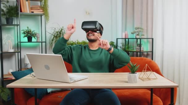Indian man using virtual reality futuristic technology VR app headset helmet to play simulation 3D 360 online video game, watching film movie at modern home apartment. Guy in goggles sitting at table - Footage, Video