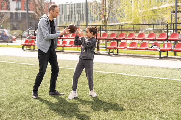 Девушка держит овальный коричневый кожаный регбийный мяч и улыбается, играя с родителями в парке. Семья и дети, концепция природы. - Фото, изображение