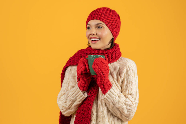 Happy young woman in winter knitted attire enjoying a cozy cup of coffee, full of festive cheer, in studio on yellow background, ideal for seasonal sale advertisements with copy space - Photo, Image