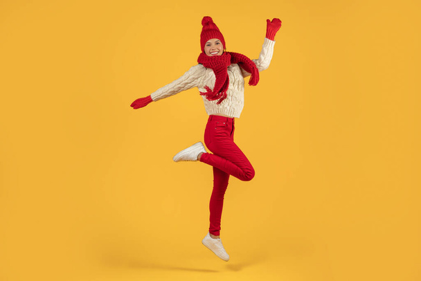 Веселая юная леди в теплой зимней одежде, прыгающая от радости, идеально подходит для рождественских и праздничных сезонов концепции, позируя в полную длину в студии на желтом фоне с копировальным пространством - Фото, изображение