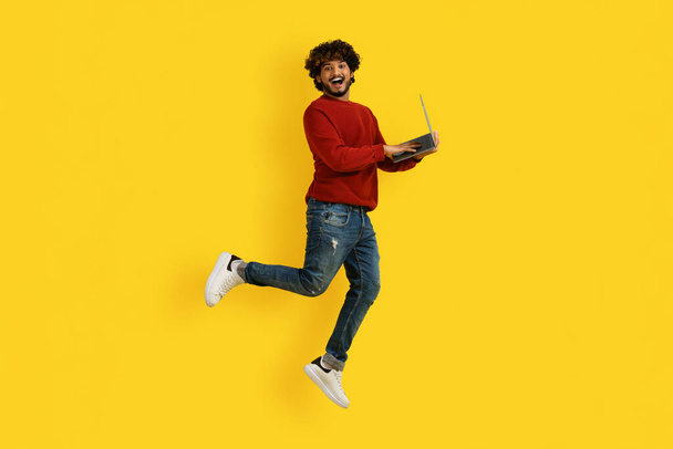 Emotional millennial indian man in casual outfit enjoying great online offer, jumping in the air with laptop computer in his hands, isolated on yellow studio background, copy space - Foto, Imagem