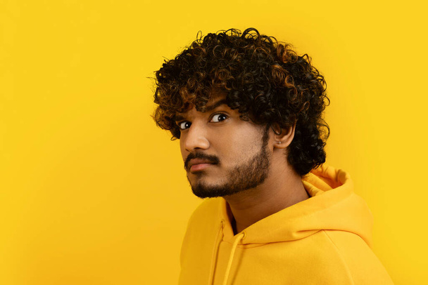 Unsure young indian bearded curly man 30s with suspicious look wearing yellow hoodie posing on camera with raising eyebrow isolated over yellow background, closeup, copy space - Valokuva, kuva