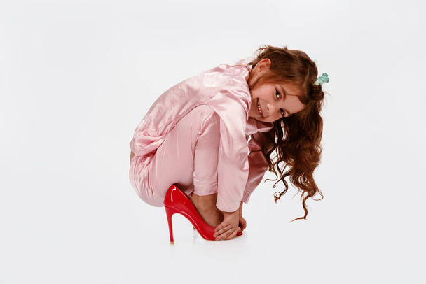 Ein nettes kleines Mädchen sitzt auf weißem Hintergrund, trägt große Schuhe mit hohen Absätzen und hält ihre Hände vor dem Mund. Große Schuhe. Kinderspaß. Große Schuhgröße. Zukünftige Dame. Hochwertiges Foto - Foto, Bild