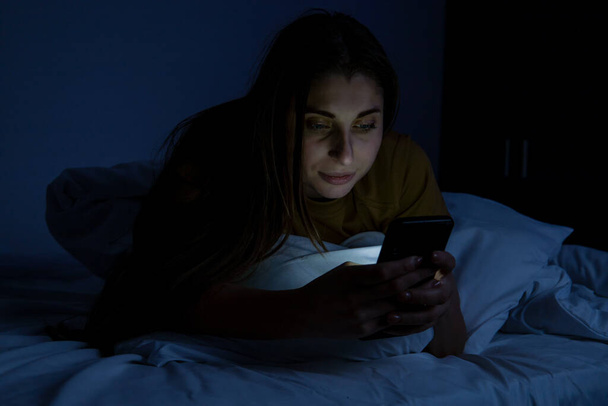Dziewczyna w łóżku z telefonem na białym łóżku w ciemnym pokoju - Zdjęcie, obraz