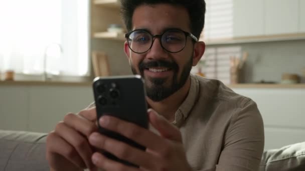 Gülümseyen Arap asıllı Hintli etnik adam erkek kullanıcı serbest çalışan ev sahibi kanepe üzerinde cep telefonu akıllı telefon dijital alışveriş mağazası uygulaması ev mutfağında online alışveriş siparişi - Video, Çekim