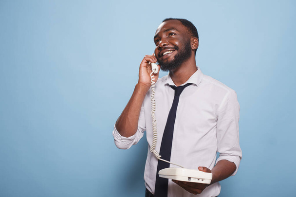 Afrykański dorosły mężczyzna wykonujący telefon na stacjonarnym. Uśmiechnięty biznesmen na analogowym telefonie, gawędzi. Współczesna osoba bawiąca się i odbierająca telefony - Zdjęcie, obraz