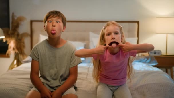 Смішні щасливі кавказькі діти хлопчик дівчинка брат сестра і сестра син дочка дражнить камеру вдома похмуро граючи разом, обдурюючи, роблячи обличчя, показуючи язик жартуючи в спальні - Кадри, відео