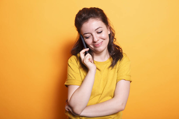 En el estudio, la mujer caucásica utiliza su teléfono inteligente para hacer una llamada telefónica a amigos. Sonriente dama morena utilizando tecnología moderna con dispositivo móvil para la comunicación telefónica remota. - Foto, imagen