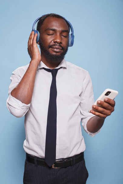 Gözleri kapalı siyah bir adamın portresi akıllı telefondan kablosuz kulaklığa kadar ses içeriğinin keyfini çıkarıyor. Genç bir ofis çalışanı, kablosuz kulaklıklardan sakinleştirici podcast dinliyor.. - Fotoğraf, Görsel