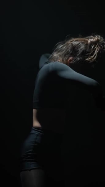 Tańcząc w słabo oświetlonym studio, oszałamiająca kobieta w czarnym ubraniu wykonuje współczesną rutynę. Wysokiej jakości materiał 4k - Materiał filmowy, wideo