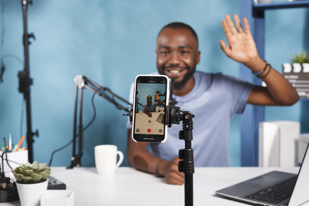 Hymyilevä serpentiini lähetykset suorana älypuhelimella jalustalla ja ammattikäyttöön tarkoitetuilla laitteilla. Afrikkalainen amerikkalainen bloggaaja tervehtii internet-kanavan yleisöä ja tallentaa videon matkapuhelimeen lähikuva - Valokuva, kuva