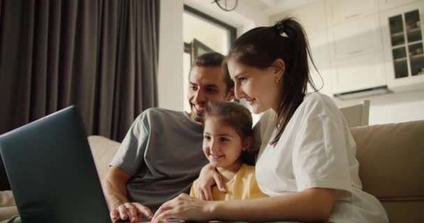 Happy family, a man with long brunette hair, his brunette girlfriend and their happy daughter in a yellow dress are sitting on a light brown something in a dark gray monitor and laughing in a modern - Filmati, video
