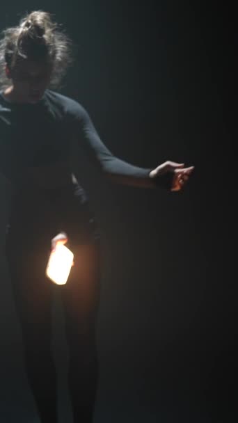 Vestida com trajes escuros, uma jovem dança com entusiasmo em uma sala mal iluminada enquanto segura uma luz. Imagens 4k de alta qualidade - Filmagem, Vídeo