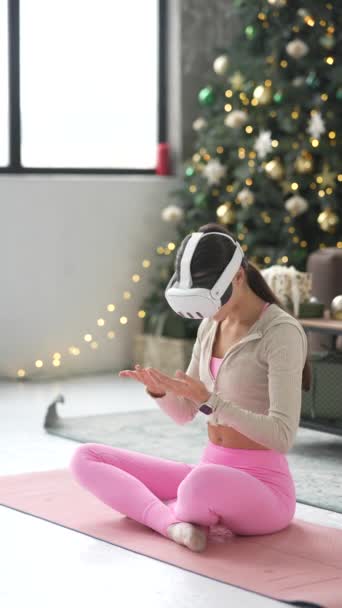 Une jeune femme mince se livre au yoga à la maison pendant les vacances de Noël, en utilisant un casque de réalité virtuelle. Images 4k de haute qualité - Séquence, vidéo