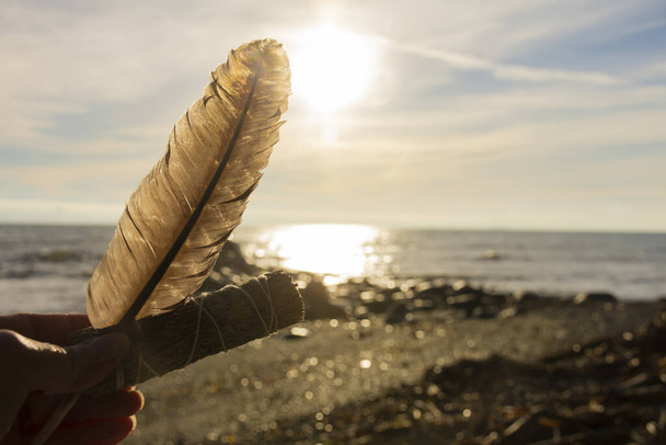 ロックの海岸で日没時に白いサージのスモッジスティックと恐怖の羽を保持する手のイメージ.  - 写真・画像