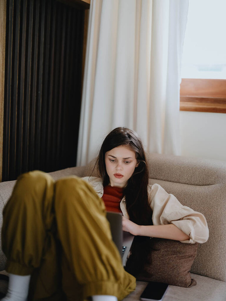 Uma mulher jaz em um sofá com um laptop e olha penosamente para a tela treinando um freelancer no trabalho, um estilo de vida real sem retocar. Foto de alta qualidade - Foto, Imagem