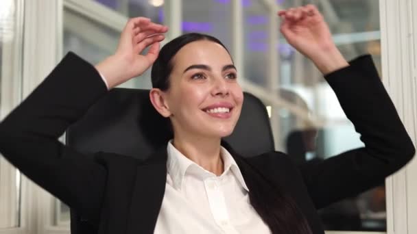 Közelkép elégedett női vállalkozó pihentető széken, és tegye a kezét a feje mögé, miközben mosolyog a szobában. Kaukázusi sikeres barna hajú fonott haj élvezi a szabadidő irodai munka. - Felvétel, videó