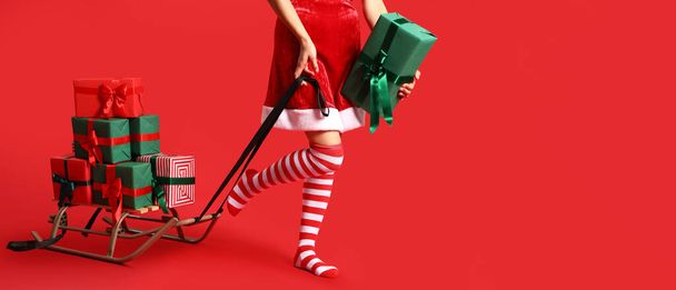 Junge Frau in Weihnachtsstrümpfen, mit Schlitten und Geschenken auf rotem Hintergrund mit Platz für Text - Foto, Bild