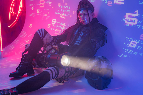 Une femme en costume noir tactique futuriste repose sur le sol, avec un casque de moto et un projecteur massif à côté d'elle, sur fond de symboles numériques projetés - Photo, image