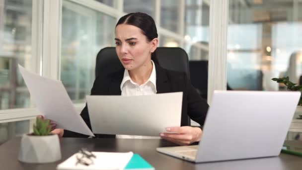 Frustrated female manager sitting in front of wireless computer and holding important documents in hands. Caucasian nervous brunette closed in black jacket looking at failed paper work in office. - Footage, Video
