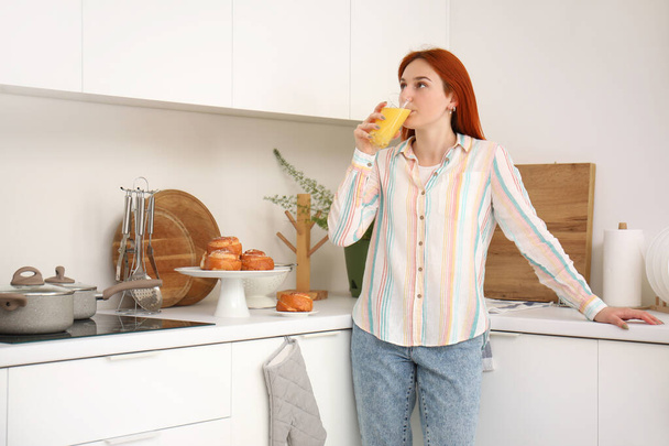 Νεαρή κοκκινομάλλα γυναίκα πίνει χυμό στην κουζίνα - Φωτογραφία, εικόνα