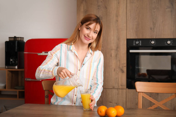 Νεαρή γυναίκα ρίχνει χυμός πορτοκαλιού στο ποτήρι στην κουζίνα - Φωτογραφία, εικόνα