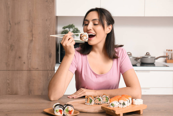 Happy young woman eating tasty sushi rolls in kitchen - Photo, Image