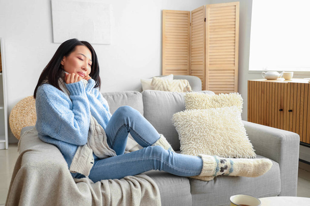 Молодая азиатка в тёплом свитере сидит дома на диване - Фото, изображение