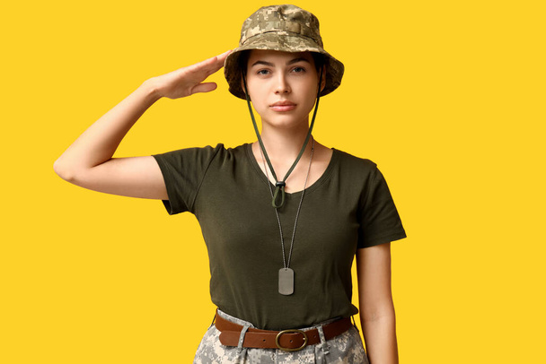 Уверенная молодая женщина-солдат в форме салютует на желтом фоне - Фото, изображение