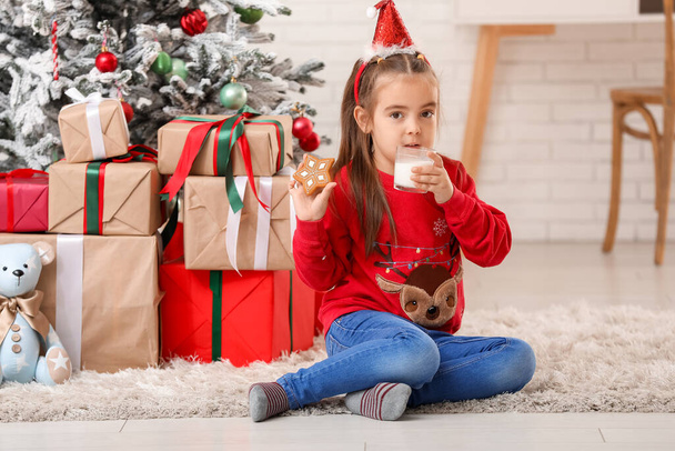 Niedliches kleines Mädchen mit Weihnachtsmütze Stirnband mit süßem Plätzchen und Glas Milch zu Hause an Heiligabend - Foto, Bild