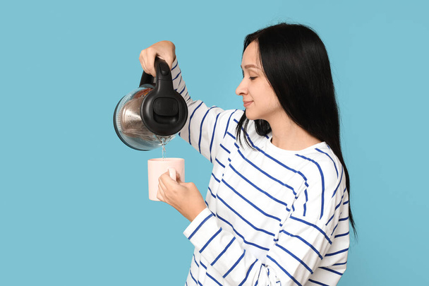 Piękna młoda kobieta wylewa ciepłą wodę z nowoczesnego czajnika elektrycznego do filiżanki na niebieskim tle - Zdjęcie, obraz
