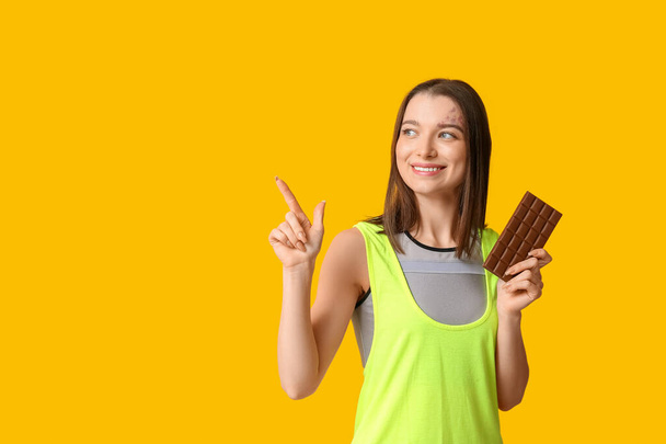 Νεαρή γυναίκα που κρατάει σοκολάτα και δείχνει κάτι σε κίτρινο φόντο - Φωτογραφία, εικόνα