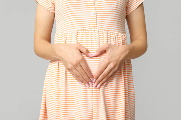 Krásná mladá těhotná žena dělat srdce s rukama na šedém pozadí, detailní záběr - Fotografie, Obrázek