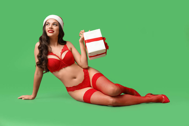Piękna młoda seksowna kobieta w bieliźnie i kapeluszu Santa z pudełkiem prezentów siedzi na zielonym tle - Zdjęcie, obraz